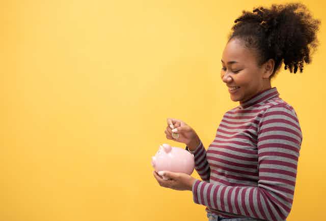 Young woman putting a coin in a piggy bank on a yellow background.