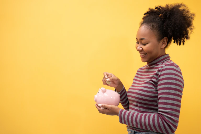 Young woman putting a coin in a piggy bank on a yellow background.