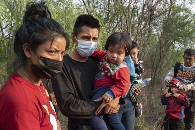 Adults, some of them wearing face masks, and children stand outdoors waiting for U.S. Border Patrol officers to pick them up.