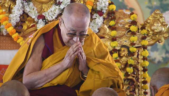 A bald man with glasses in red and gold robes sits on an ornate golden-colored chair.