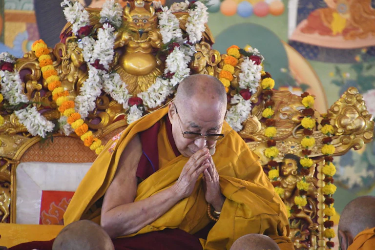 A bald man with glasses in red and gold robes sits on an ornate golden-colored chair.