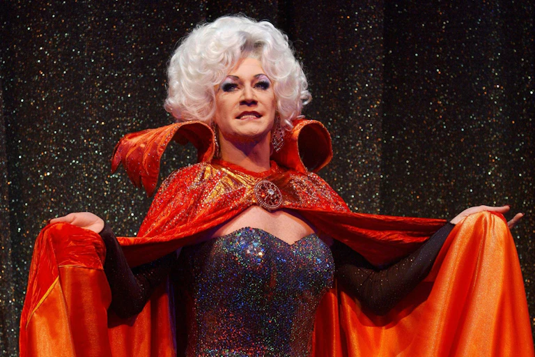 Drag queen Lily Savage dressed as the wicked queen in the pantomime Snow White, dressed in a sparkly black dress and huge orange cape.