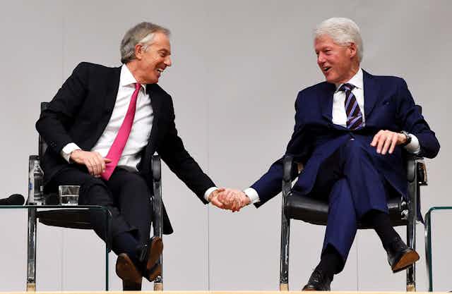 Tony Blair and Bill Clinton laughing and holding hands on stage at an event.