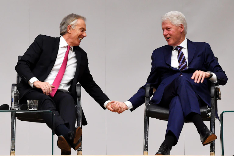 Tony Blair and Bill Clinton laughing and holding hands on stage at an event.