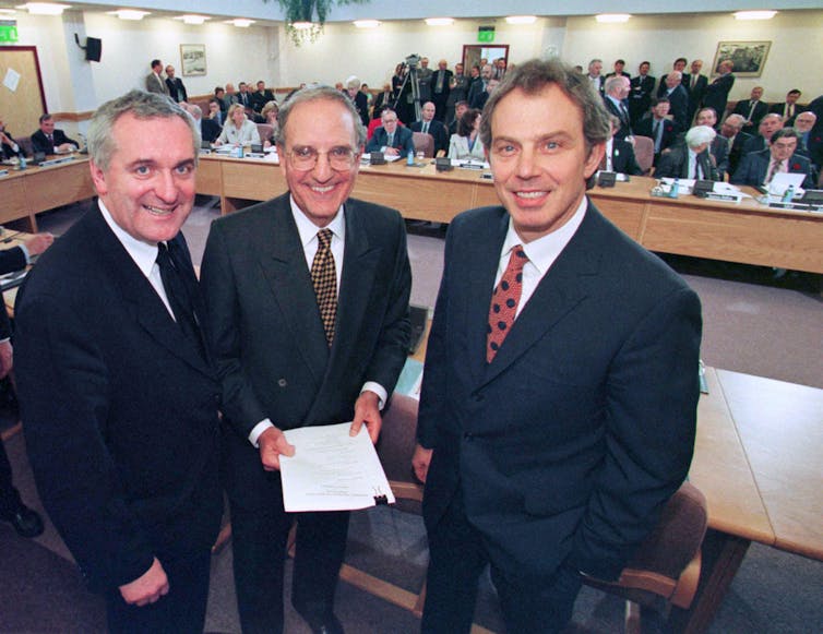 Bertie Ahern, George Mitchell and Tony Blair standing for a photo. Mitchell is holding a copy of the Good Friday Agreement.