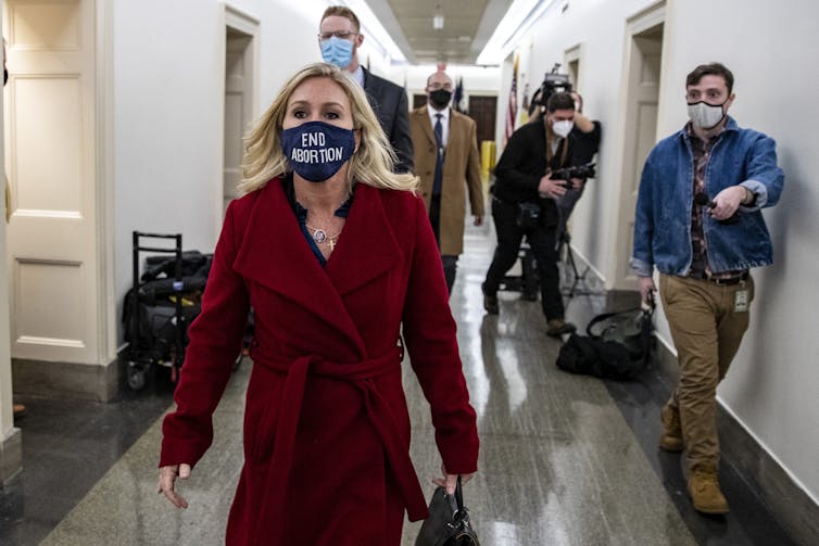 A woman wears a red coat and a face mask that says 'end abortion.' She walks down a hallway with men behind her, also wearing masks.
