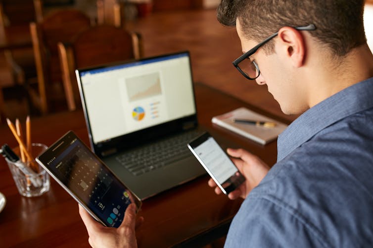 a man holds two smartphones in his hand while sitting in front of a laptop showing charts on its screen