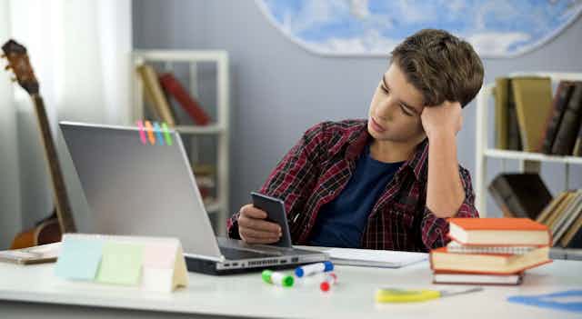 a teenaged boy sits at a desk with a laptop. he's holding a smartphone and a pile of four books rests next to him