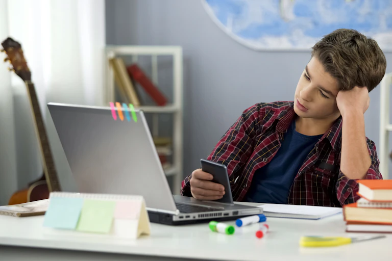a teenaged boy sits at a desk with a laptop. he's holding a smartphone and a pile of four books rests next to him
