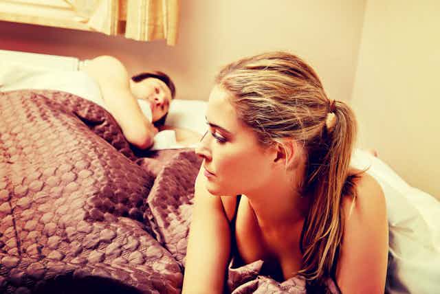 Worried woman sitting on edge of bed while partner sleeps