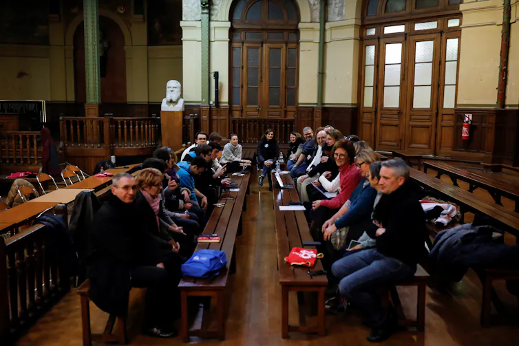 A dozen of people sit on benches around a table, in a public building