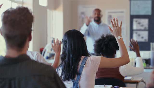 A Black male teacher stands at the front of a high school class as students raise their hands. The image of the teacher is distorted.