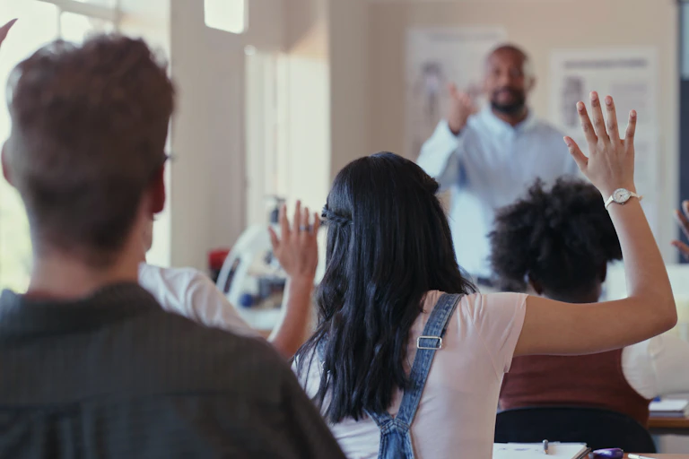 A Black male teacher stands at the front of a high school class as students raise their hands. The image of the teacher is distorted.