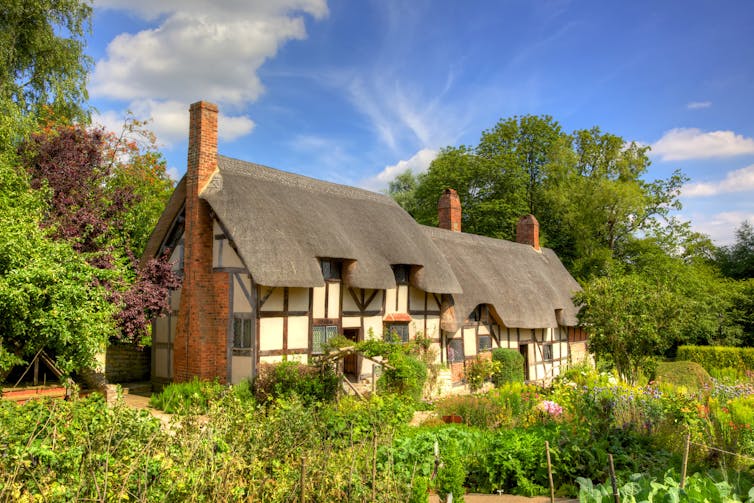 A thatched English cottage with white walls surrounded by greenery, photographed under blue skies.