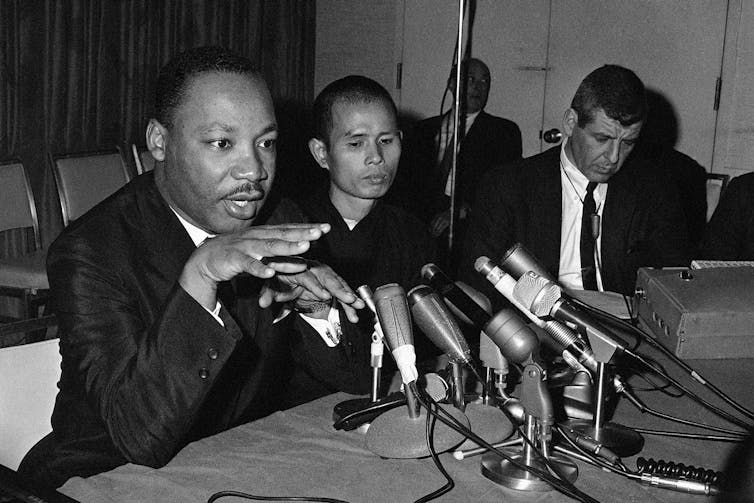 Two men, one in a suit and one in a monk's robe, speak into microphones on a table.