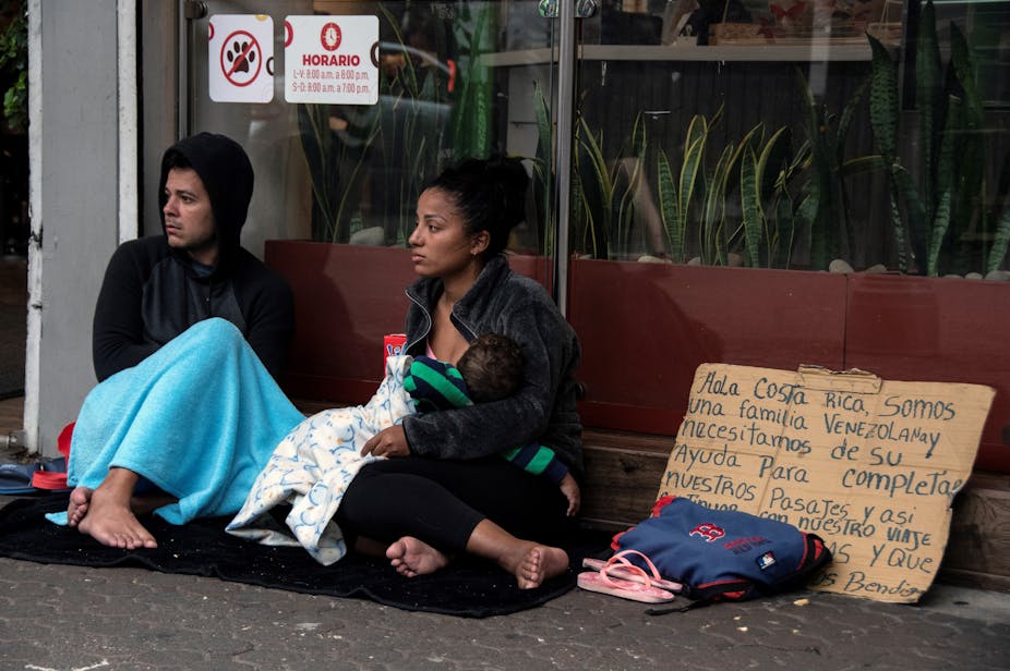 Un homme et une femme assis en train de mendier à côté d'une pancarte en espagnol expliquant qu'ils viennent du Venezuela et se dirigent vers les États-Unis