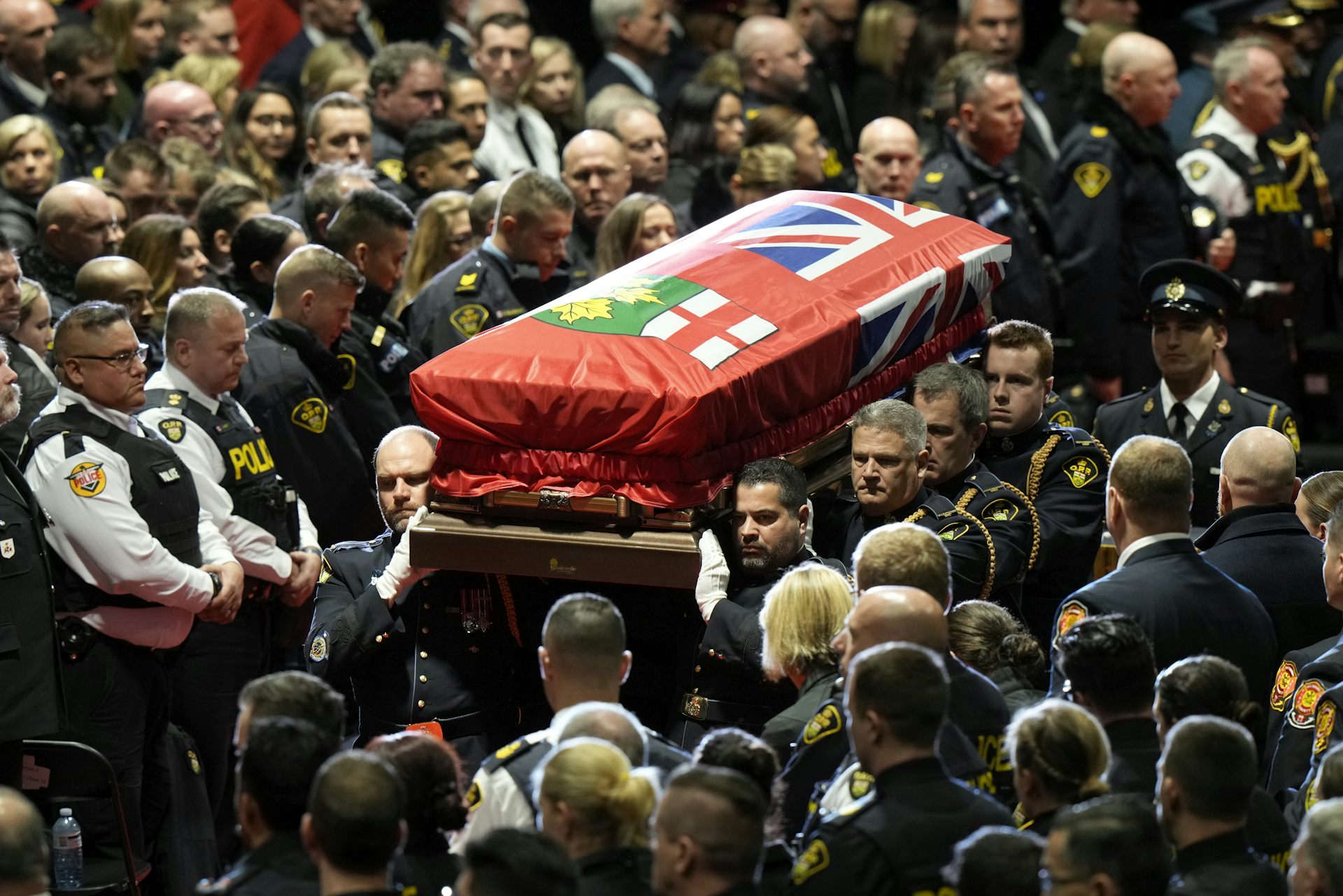Pallbearers carry a coffin draped with a mostly red flag past a large group of mourners.