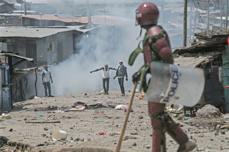 A man wearing a body armour and helmet, and carrying a baton and police shield looks at three men who have a plume of smoke behind them, and debris in front of them