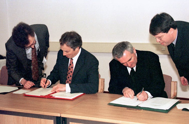 Tony Blair and Bertie Ahern sitting at a table signing copies of the Good Friday Agreement with staff directing them.