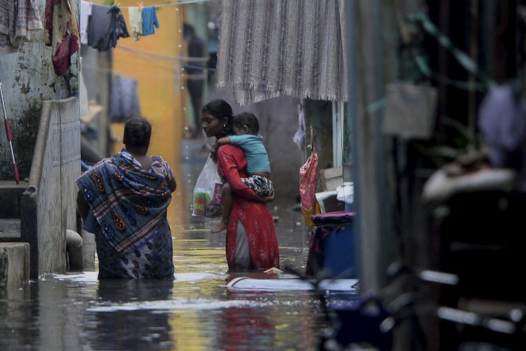 A brown skinned woman wears a red dress and carries a child on her back while walking through water up to her knees.