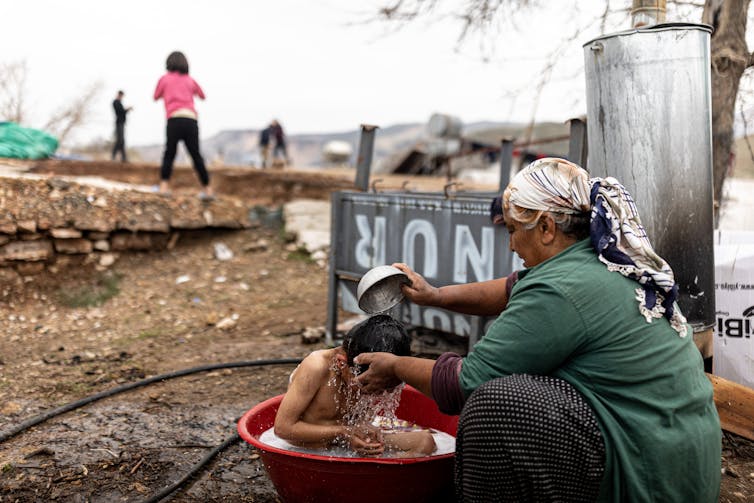 A woman in a headscarf and a green shirt pours a bowl of water over a child's head. The child sits in a red bucket of water.