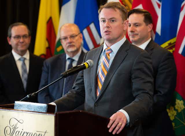 A man with dark blond hair in a grey suit and striped tiespeaks into a microphone with three men standing behind him in front of a row of flags.