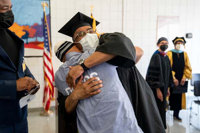 A man wearing a black graduation cap and gown hugs a man wearing a blue prison uniform
