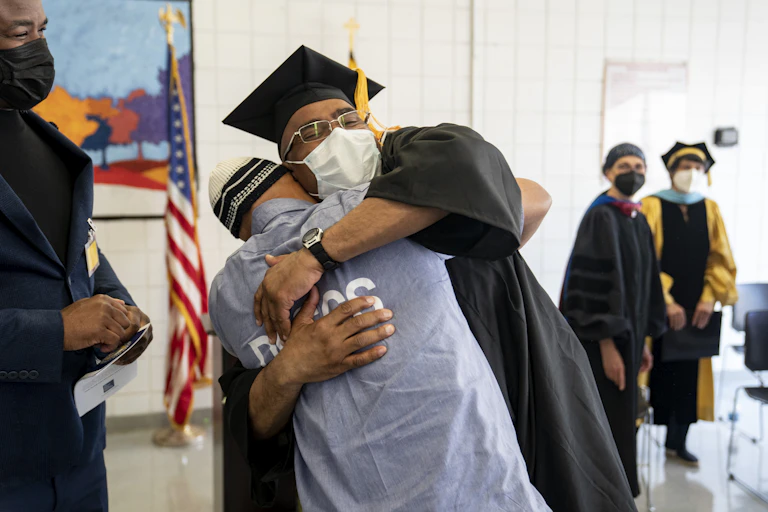 A man wearing a black graduation cap and gown hugs a man wearing a blue prison uniform