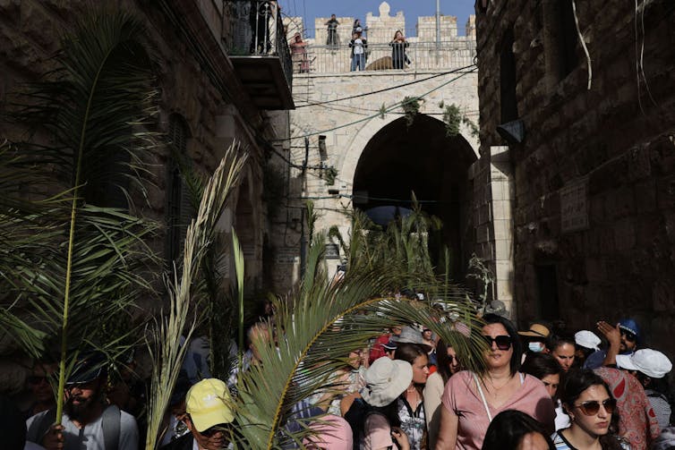 A crowd of people some of them holding tall palm branches walk through a narrow street in an ancient city