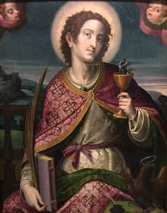 A painting shows a male saint with a halo holding a cup with a small dragon in it and a palm leaf