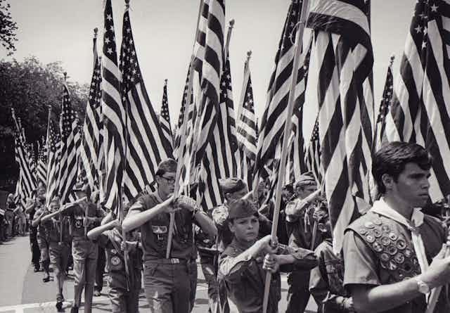 Black and white photo of Boy Scouts marching while holding American flags aloft
