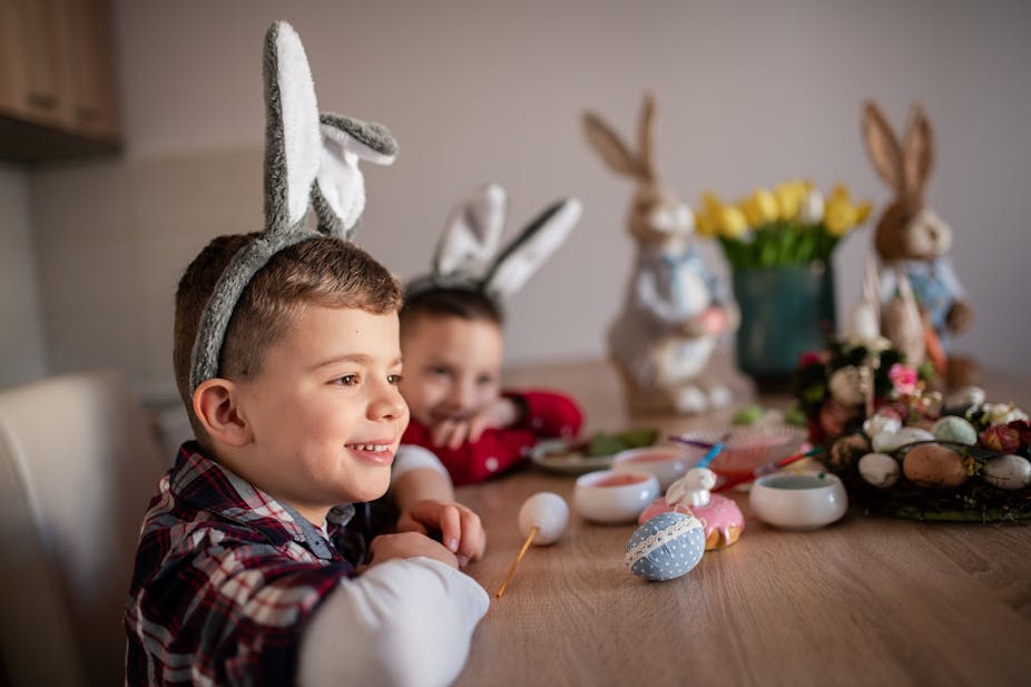 Dos niños con orejas de conejo sobrepuestas sonríen y pintan huevos de Pascua. 