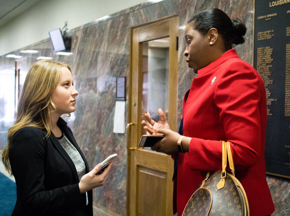 A person with long blonde hair wearing a dark blazer holds a cellphone while talking with a person with black hair pulled back in a bun wearing a red jacket.