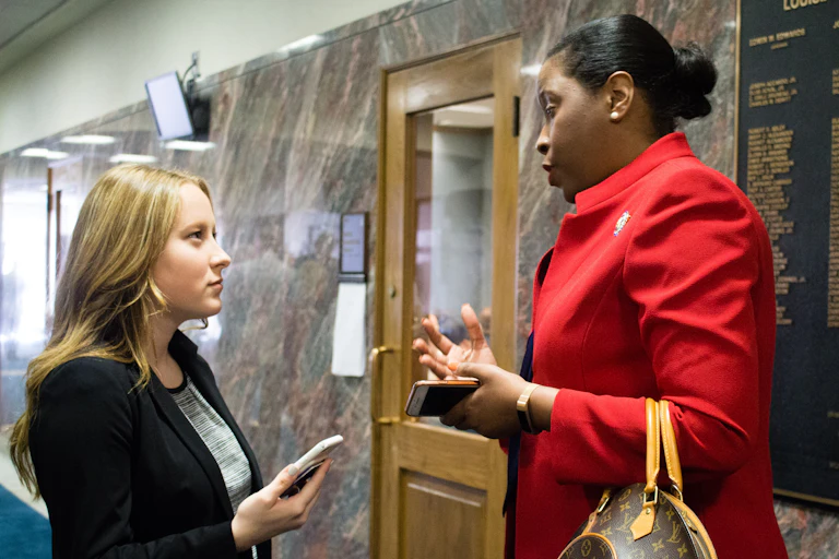 A person with long blonde hair wearing a dark blazer holds a cellphone while talking with a person with black hair pulled back in a bun wearing a red jacket.