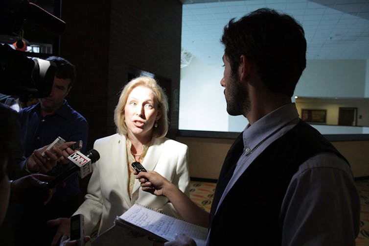 A person lit brightly speaks into a group of microphones, one of which is held by a person visible but in shadow.