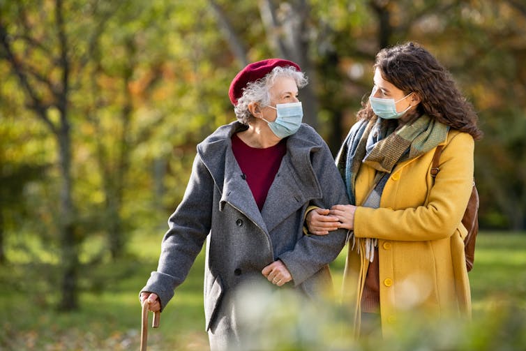 An older woman walking outdoors with a younger woman who has her arm through the older woman's. Both are wearing face masks.