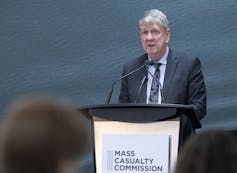 A grey-haired man speaks into a microphone. A sign that reads Mass Casualty Commission is on the front of the podium.