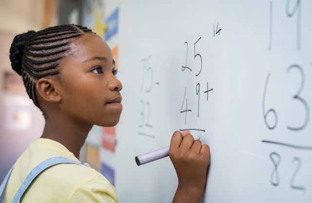 A young school girl writes a math problem on a whiteboard using a magic marker.