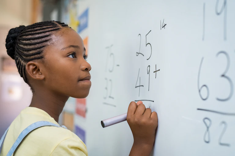 A young school girl writes a math problem on a whiteboard using a magic marker.