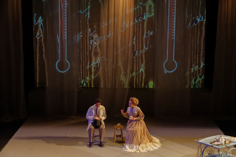 Two actors seen sitting on a stage.
