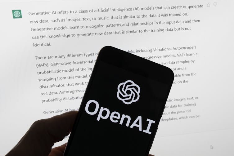 A hand holds a phone with the OpenAI logo displayed on it in front of a computer screen with a ChaptGPT discussion on it