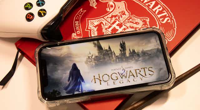 A phone showing Hogwarts Legacy is seen on top of a red book that says Hogwarts.