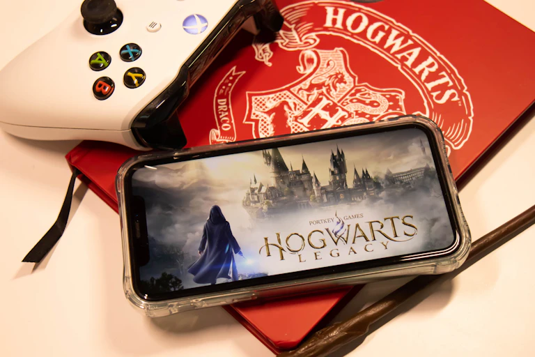 A phone showing Hogwarts Legacy is seen on top of a red book that says Hogwarts.