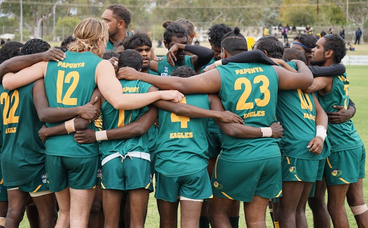 A group of First Nations people in footy guernseys are in a huddle on a football field.