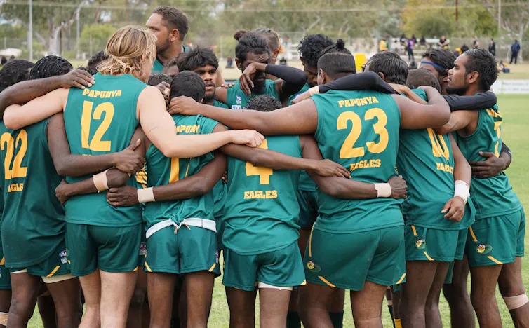 A group of First Nations people in footy guernseys are in a huddle on a football field.
