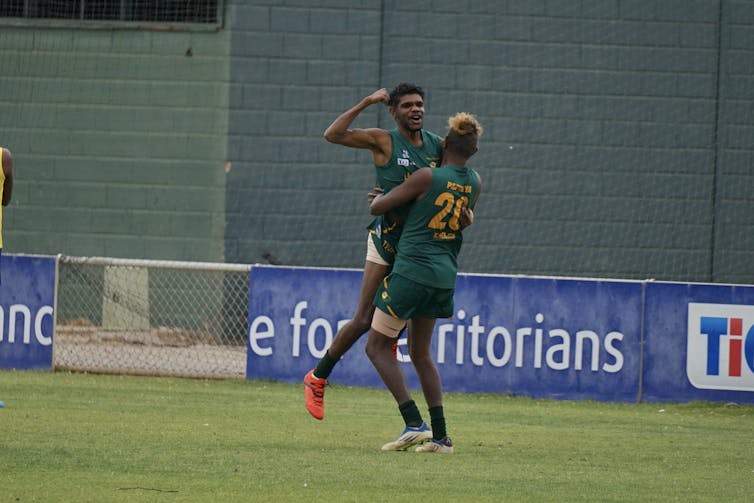 Two First Nations people are playing football on an oval. One lifts up the other in celebration. The person being lifted has a big smile with their fist in the air.