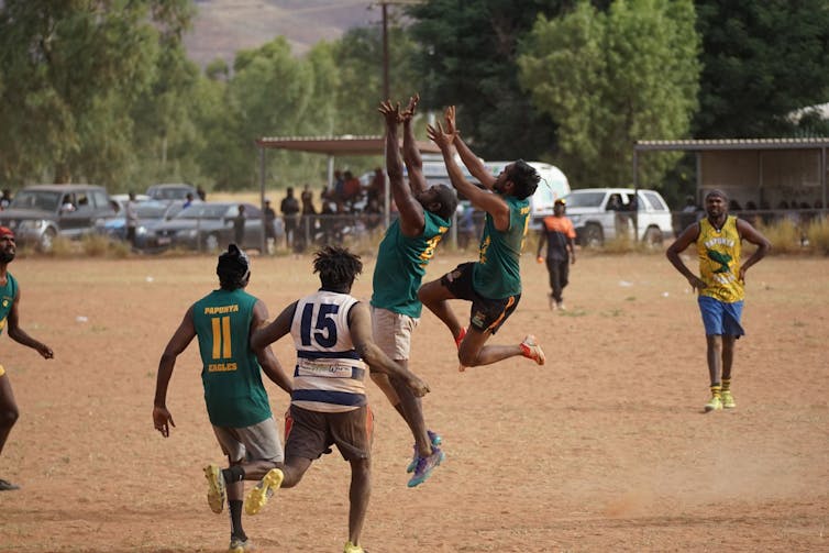 A group of First Nations footballers play football on a dusty oval. Two of them are mid-leap to catch the ball.