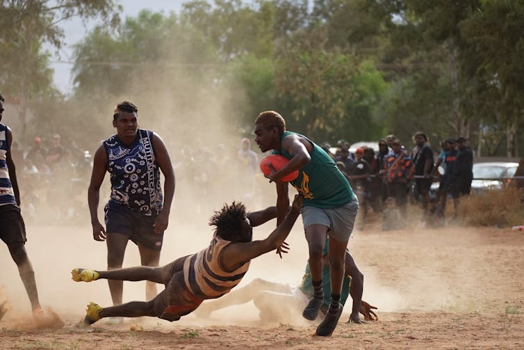 First Nations football players play on a dusty field.