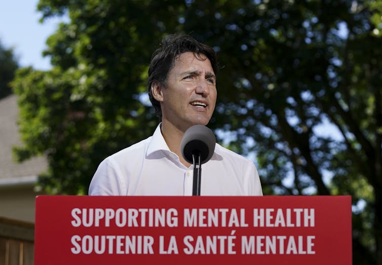 Prime Minister Justin Trudeau wearing a white shirt speaking at a podium behind a sign reading 'Supporting Mental Health'