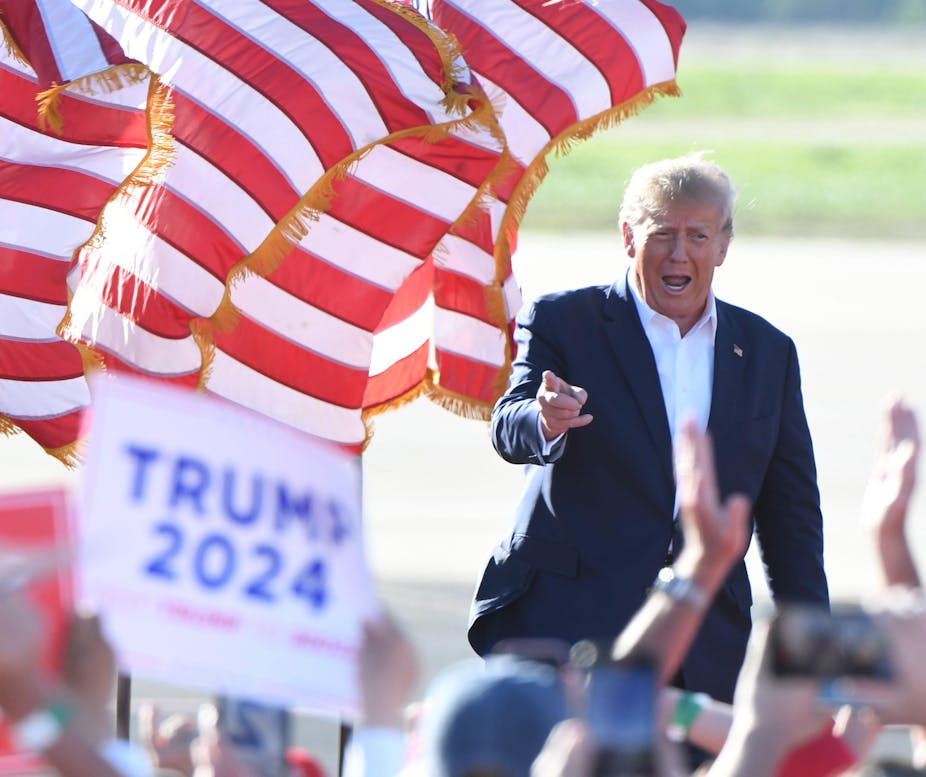Donald Trump wearing suit jacket and white shirt in front of red and white striped banner.